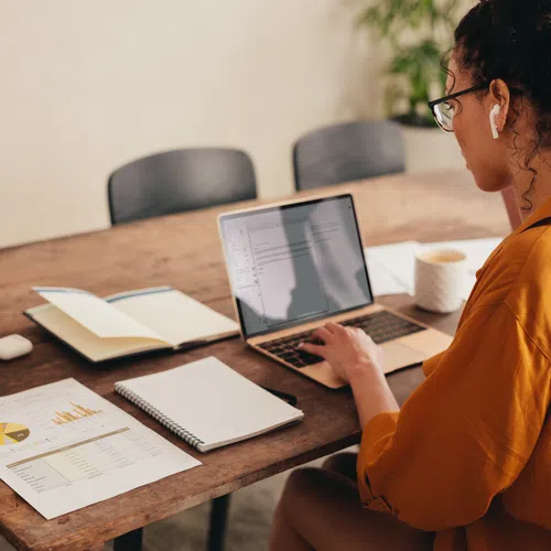 Woman in orange shirt working on laptop with documents and notebooks on wooden table in office.