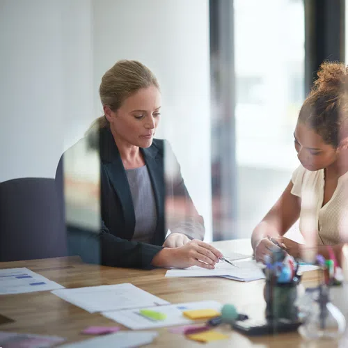 Two professional women discussing documents at a desk in a modern office setting.