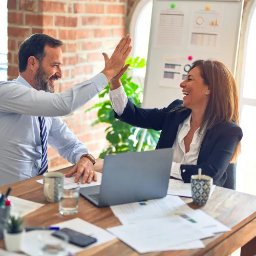 Businessman and businesswoman in office smiling and giving a high five over a laptop and paperwork