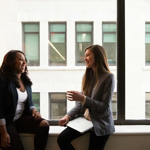 Two professional women chatting and laughing by a large window in a modern office setting