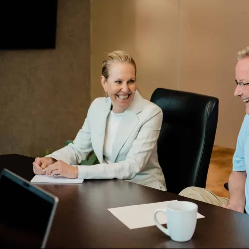 Two colleagues smiling and chatting at a conference table with coffee and documents in a bright office.