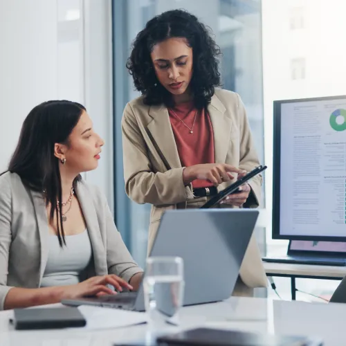 Two professional women discussing data charts on a tablet and computer in a modern office setting