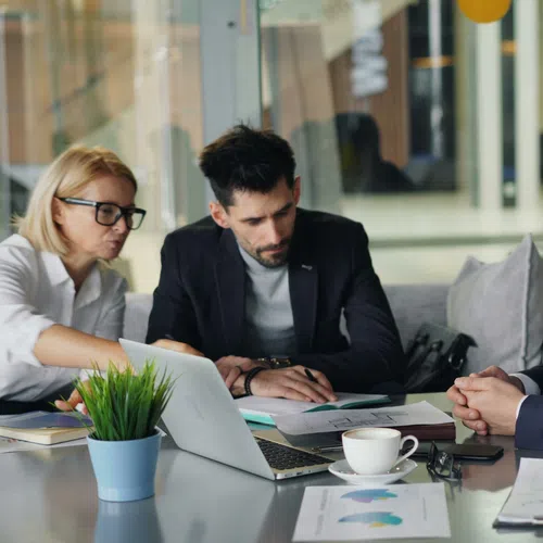 Three professionals in business attire discussing documents in an office meeting with laptop and charts on the table