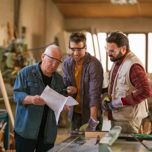 Three men in safety glasses discuss woodworking plans together in a workshop filled with lumber and tools.