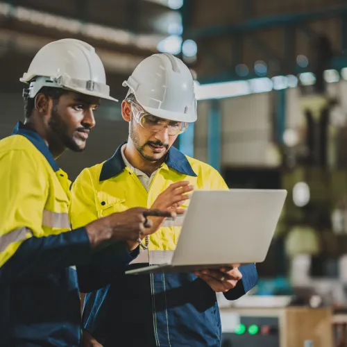 Two construction workers in safety gear using a laptop in an industrial warehouse setting.