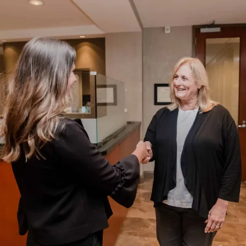 Two professional women shaking hands in a modern office lobby, smiling and greeting each other warmly.