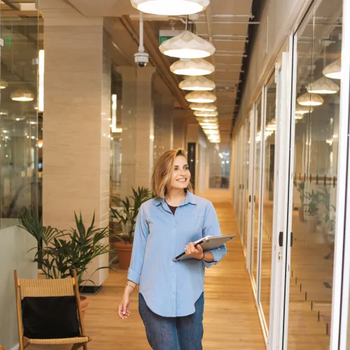 Smiling woman walking in modern office hallway holding a laptop, surrounded by plants and glass walls.