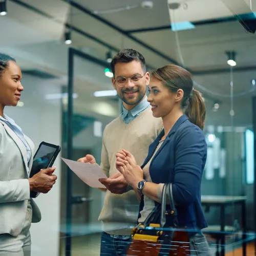 Three professionals engage in a discussion inside a modern office with papers and a tablet.