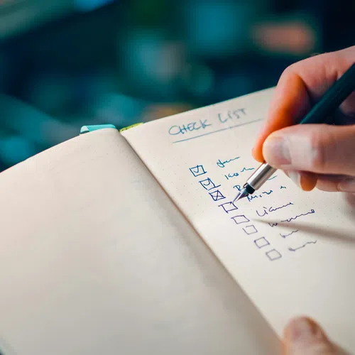 Hand holding a pen checking off items on a handwritten checklist in a notebook with a laptop keyboard in the background