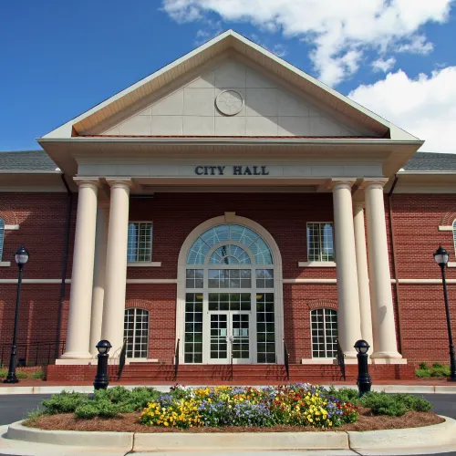 Red brick city hall building with tall white columns, arched windows, and flowerbed in front under a blue sky.