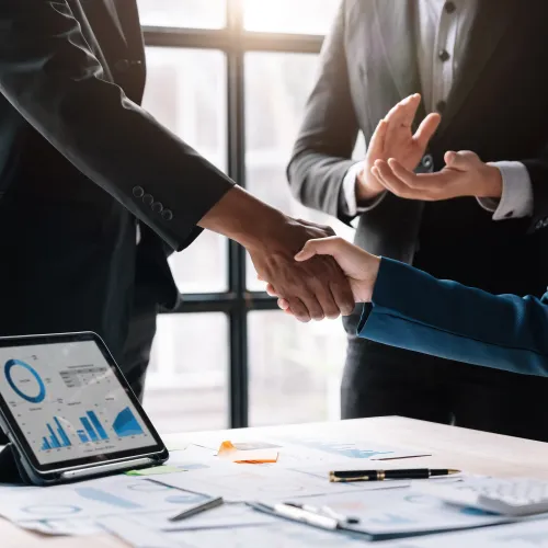 Business professionals shaking hands in office with financial charts and tablet on the table confirming a deal.