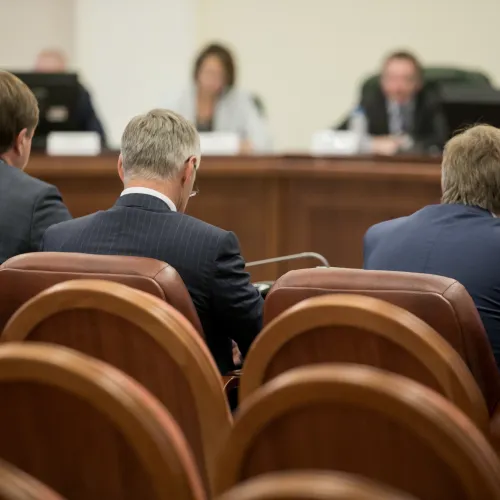 Three men in suits sitting in a conference room with blurred panelists in the background during a meeting.