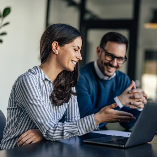 Two colleagues smiling and discussing work while looking at a laptop in a modern office setting.
