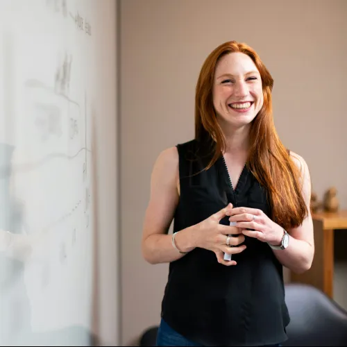 Smiling red-haired woman in black sleeveless top stands by a whiteboard with diagrams, reflecting on the glass surface.