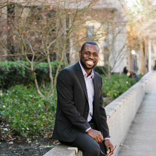 Smiling man in black suit sitting on urban stone ledge surrounded by greenery and city buildings.