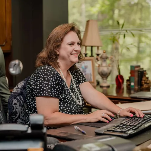 Woman in a black and white patterned blouse smiling while typing on a keyboard at a desk with a computer and office decor.
