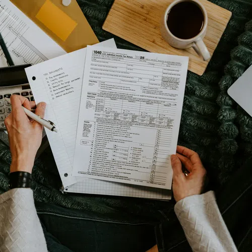 Person filling out tax forms with calculator, notepad, laptop, and coffee cup on a green blanket.