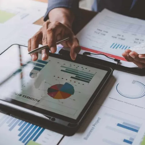 Person analyzing business charts and graphs on a tablet and printed reports with a pen in hand.
