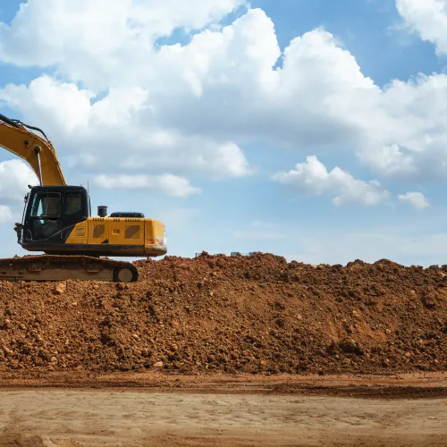 Yellow excavator on large dirt mound under a blue sky with white clouds at a construction site.