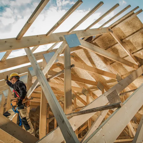 Construction worker wearing safety gear working on wooden roof framing under a blue sky with clouds.