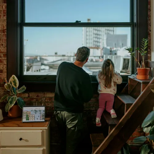 Man and young girl looking out large window in cozy urban apartment with plants and wooden stairs.