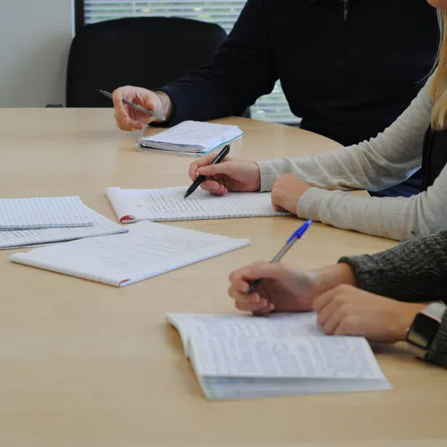 People writing notes in notebooks during a meeting at a wooden table in an office setting.