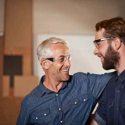 Two men wearing safety glasses smiling and embracing each other in a woodworking workshop.