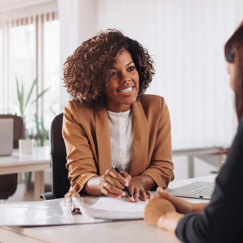 Two women having a professional conversation in a modern bright office with documents and laptop on desk.