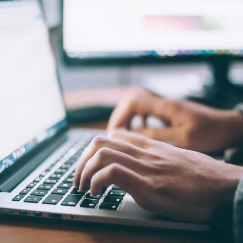 Person typing on a laptop keyboard with dual monitors in the background in a workspace setting