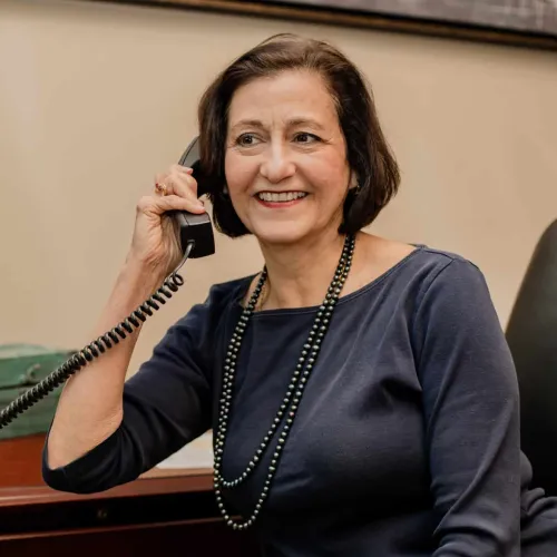 Smiling mature woman talking on a corded office phone at her desk wearing a navy blue top and black beaded necklace