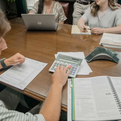 Group of professionals at wooden table using calculator, laptop, and documents during office meeting.