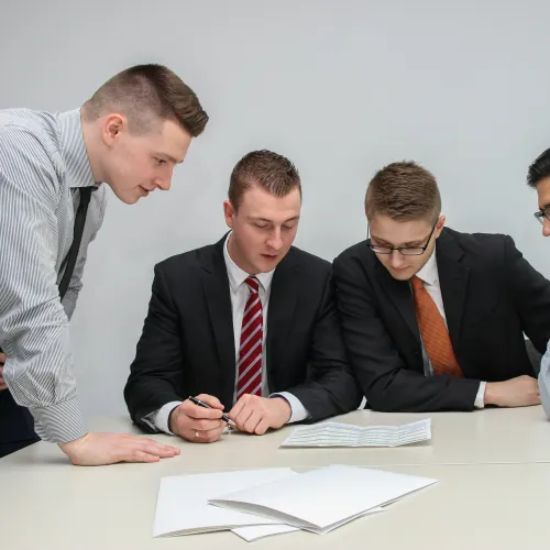 Four young professionals in suits and ties engaged in a collaborative meeting around a table with documents.