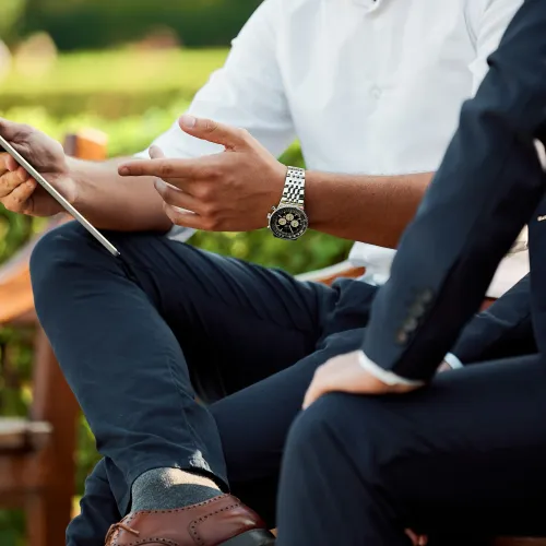 Two men in business attire sitting on a bench discussing while holding a tablet outdoors in a park.