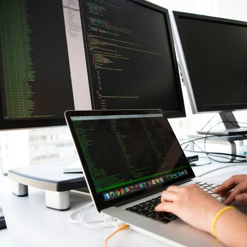 Person typing code on laptop with dual monitors displaying programming scripts in a modern office setup