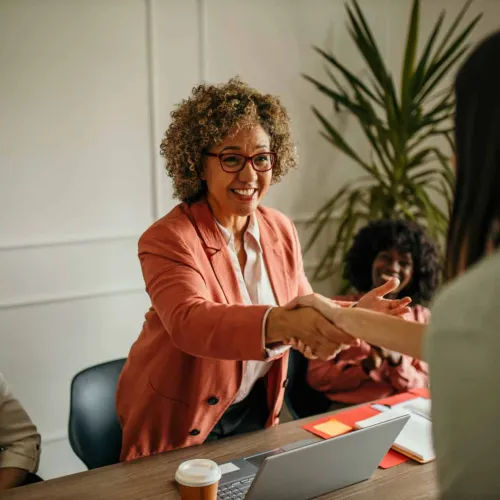 Smiling woman in a pink blazer shaking hands with another person during a business meeting at a desk.