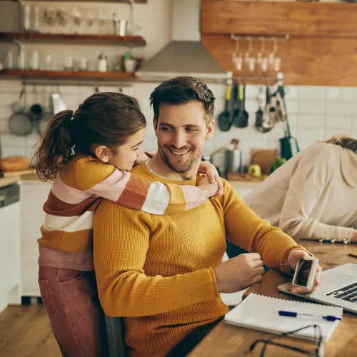 Father embraces daughter while working on laptop in cozy kitchen with mother and younger child nearby
