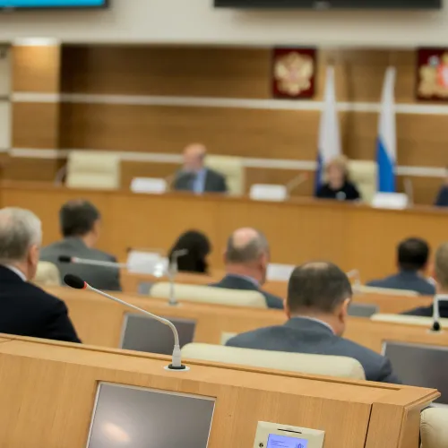 Conference room with officials seated, microphones, and flag emblems in a formal government setting.