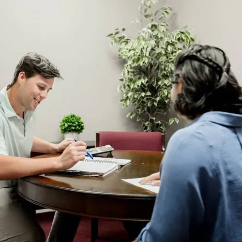 Two people sitting at a round table discussing notes with a calculator and plant in background