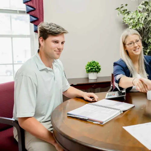 Two professionals shaking hands across a table during a business meeting with documents and calculator.