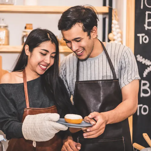 Smiling couple in aprons baking together in a cozy kitchen holding a plate with a freshly baked pastry.