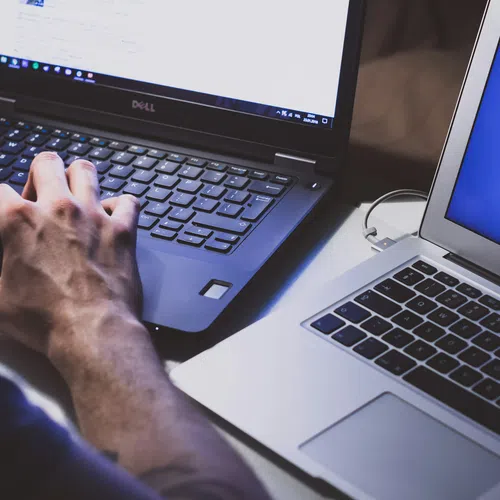 Person typing on Dell laptop next to an open MacBook Air on a desk with screens showing software windows