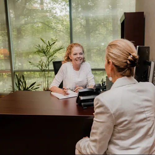 Two women in a professional office setting engaged in a friendly conversation at a wooden desk.