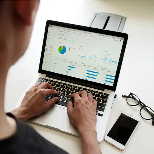 Person analyzing data charts and graphs on a laptop with smartphone and glasses on desk beside them