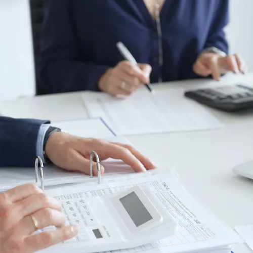 Two business professionals working with documents and calculator on office desk with laptop and files.