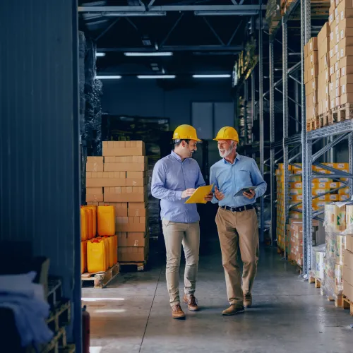 Two warehouse workers wearing yellow helmets discussing inventory with tablet and clipboard in storage aisle.