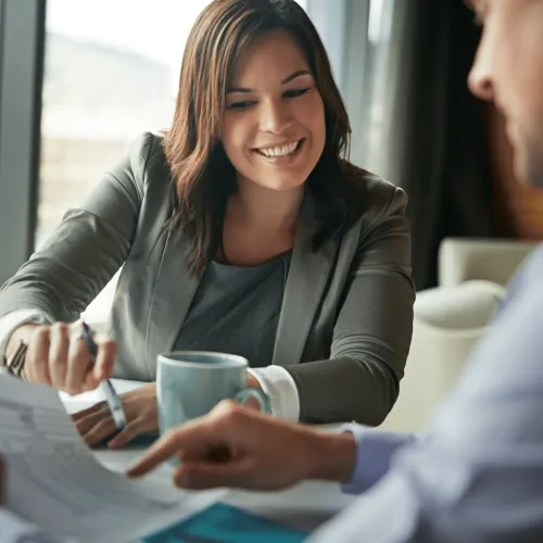 Businesswoman smiling and discussing documents with a male colleague in an office setting over coffee.