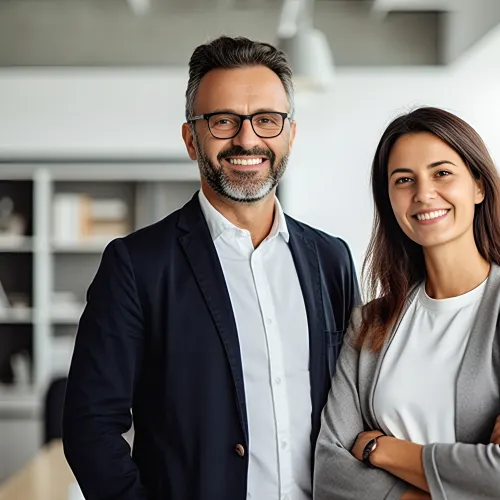 Confident man and woman in business attire smiling in modern office environment with natural light.
