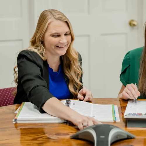 Two women smiling and working together at a table with documents and a conference phone