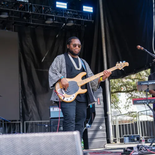 Musician playing electric bass guitar onstage with keyboardist performing outdoors during daytime concert.