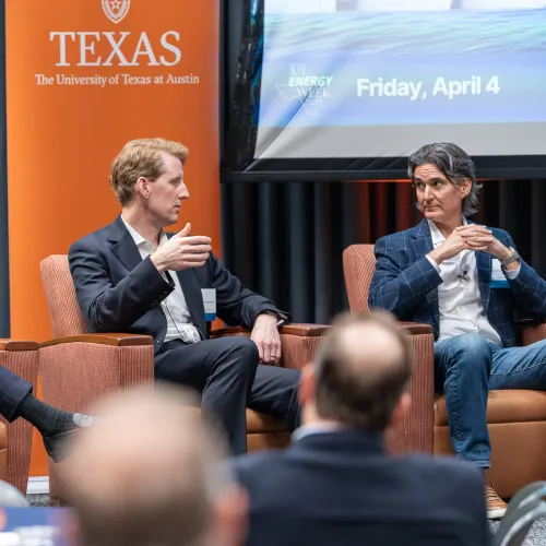 Three men in business attire engaged in a panel discussion at University of Texas event with branded banners behind them